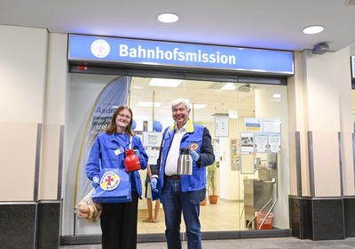 Zwei Mitarbeiter der Bahnhofsmission stehen vor dem Eingang mit einem großen Schild. Die Frau hält eine rote Kanne und eine Tüte mit Brötchen, der Mann hält eine silberne Kanne. Beide tragen blaue Jacken und lächeln freundlich.
