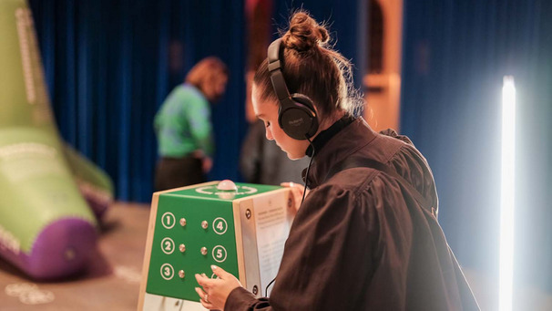 Eine Frau steht in einer Ausstellung, trägt Kopfhörer und lässt sich einen Text vorlesen