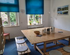 Ein helles, freundliches Zimmer mit einem großen Holztisch in der Mitte. Um den Tisch sind einfache Bänke mit Sitzkissen angeordnet. An einem Fenster stehen Blumen und es gibt eine Stehlampe. Blaues Licht fällt durch die Fenster mit Vorhängen.