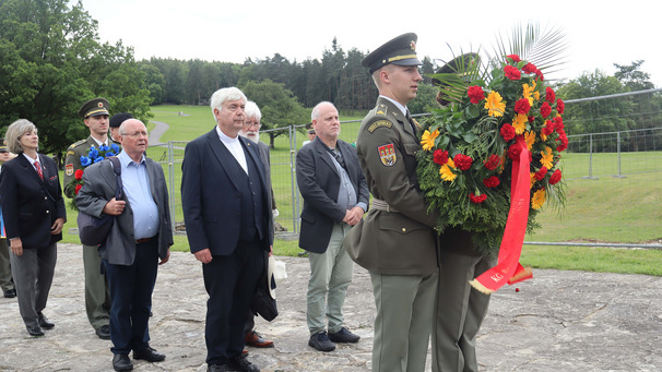 Eine Gruppe von Menschen steht auf einer Wiese. Ein Soldat präsentiert einen Kranz aus bunten Blumen, während die anderen Teilnehmer aufmerksam schauen. Der Kranz ist mit roten, gelben und grünen Pflanzen gestaltet. 