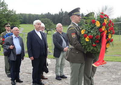 Eine Gruppe von Menschen steht auf einer Wiese. Ein Soldat präsentiert einen Kranz aus bunten Blumen, während die anderen Teilnehmer aufmerksam schauen. Der Kranz ist mit roten, gelben und grünen Pflanzen gestaltet. 