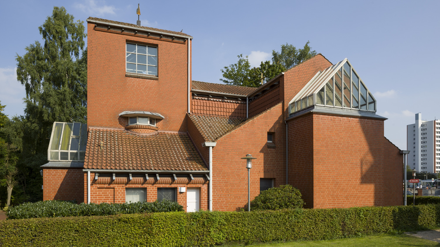 Ein modernes, zweigeschossiges Gebäude aus roten Ziegeln. Es hat ein auffälliges, geneigtes Dach mit einer Glasstruktur und große Fenster. Vor dem Gebäude befindet sich eine gepflegte Hecke und ein kleiner Weg. Im Hintergrund sind Bäume und ein urbanes Umfeld sichtbar.