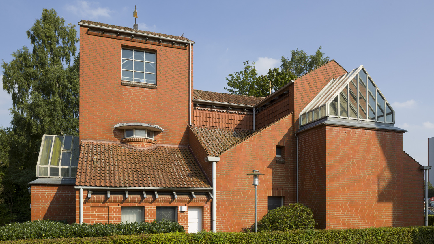 Ein modernes, zweigeschossiges Gebäude aus roten Ziegeln. Es hat ein auffälliges, geneigtes Dach mit einer Glasstruktur und große Fenster. Vor dem Gebäude befindet sich eine gepflegte Hecke und ein kleiner Weg. Im Hintergrund sind Bäume und ein urbanes Umfeld sichtbar.