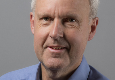 Ein Mann mit grauen Haaren und einem freundlichen Lächeln trägt ein hellblaues Hemd. Er steht vor einem neutralen grauen Hintergrund. Sein Blick ist direkt in die Kamera gerichtet, was Vertrauen und Offenheit vermittelt.