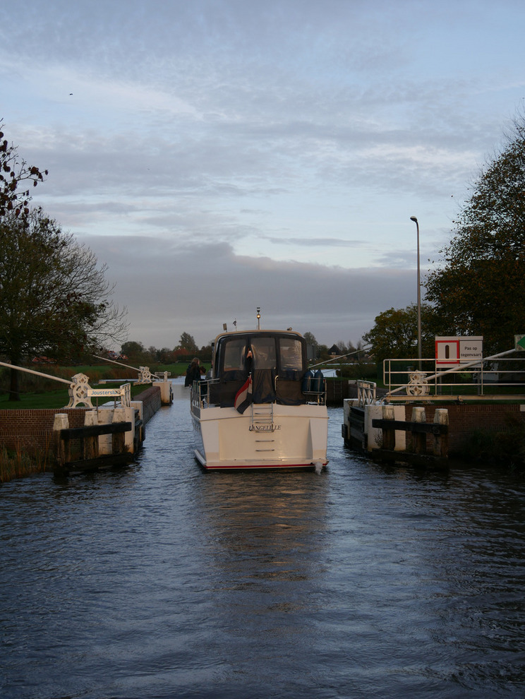 Wouter von hinten Ein Boot fährt durch eine schmale Schleuse, links und rechts vom Boot sind die geöffneten Schleusentore. Das Boot ist von hinten zu sehen. Im Hintergrund sind Bäume und ein bewölkter Himmel sichtbar.