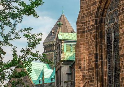 Detailansicht von Dachspitzen einer historischen Kirche. Die Dächer sind mit grünem Kupfer eingedeckt und zeigen verschiedene architektonische Stile. Im Hintergrund sind Wolken am Himmel sichtbar, während Bäume im Vordergrund leicht ins Bild ragen.