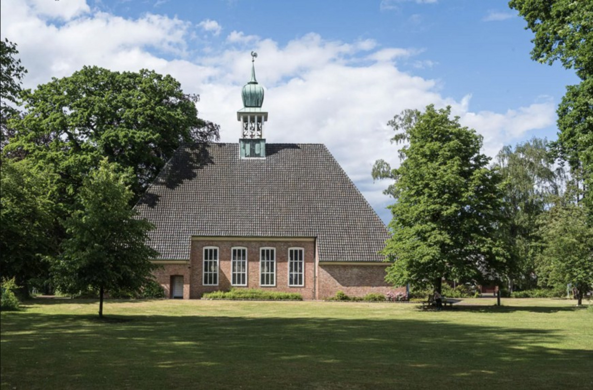 Ein historisches Gebäude mit einem schrägen Dach und einem kleinen Turm steht inmitten einer grünen Landschaft. Bäume umgeben die Architektur, und der Himmel ist teilweise bewölkt. Die Fassade ist aus Ziegelsteinen und hat große Fenster.