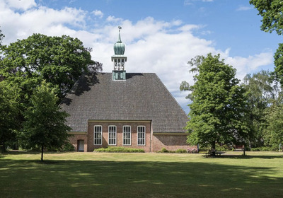 Ein historisches Gebäude mit einem schrägen Dach und einem kleinen Turm steht inmitten einer grünen Landschaft. Bäume umgeben die Architektur, und der Himmel ist teilweise bewölkt. Die Fassade ist aus Ziegelsteinen und hat große Fenster.