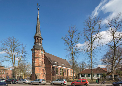 Eine historische Kirche mit einem hohen, spitzen Turm und einer Turmuhr steht vor einem klaren blauen Himmel. Umgeben von kahlen Bäumen und parkenden Fahrzeugen auf einer Straße. Der Himmel ist klar mit einigen Wolken.