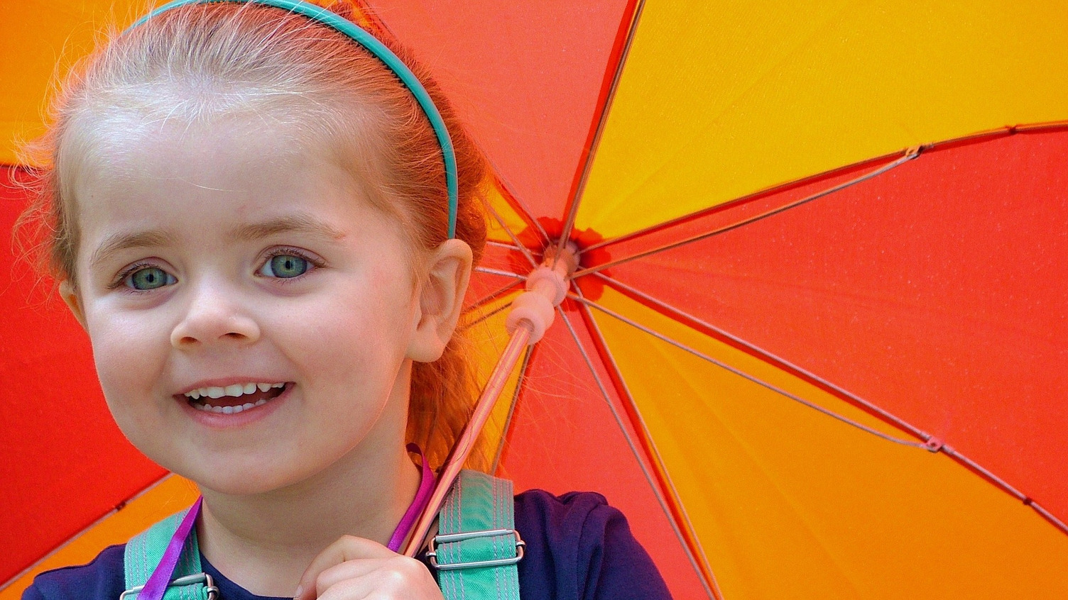 Ein lächelndes Mädchen mit blonden Haaren hält einen bunten Regenschirm in orange und gelb. Sie trägt ein dunkelblaues Oberteil und einen grünen Latz. 