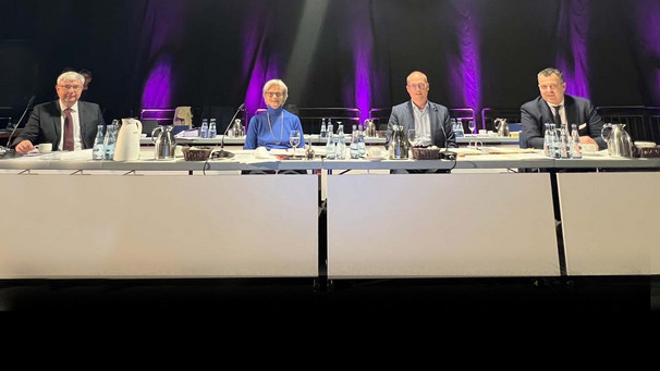 Auf dem Foto ist ein Podium mit dem Vorstand des Kirchenausschuses zu sehen. V.l.: Schriftführer Pastor Bernd Kuschnerus, Präsidentin Edda Bosse, Vizepräsident Martin Franzius, Schatzmeister Oliver Gampper.