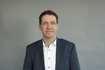 Ein Mann mittleren Alters steht vor einem grauen Hintergrund. Er trägt einen dunklen Anzug mit einem weißen Hemd und blickt freundlich in die Kamera. Seine Haare sind kurz und dunkel. Die Pose strahlt Selbstbewusstsein und Professionalität aus.