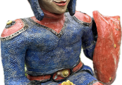 Eine farbenfrohe Figur eines Ritters in blauer und roter Kleidung, der lächelt. Er hält eine Krone in der linken Hand und hat ein Schild an seiner rechten Seite. Die Statue hat detailreiche Gesichtszüge und eine markante Kopfbedeckung.