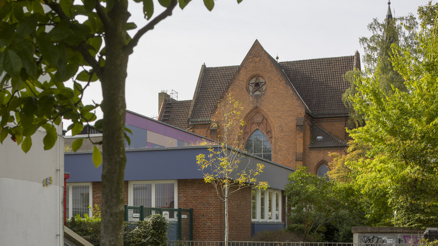 Ein rotes Backsteingebäude mit einem hohen, schrägen Dach und einem großen Fenster ist im Hintergrund sichtbar. Im Vordergrund stehen einige Bäume und ein kleiner Gartenbereich. Der Himmel ist klar, und die Umgebung wirkt ruhig und gepflegt.