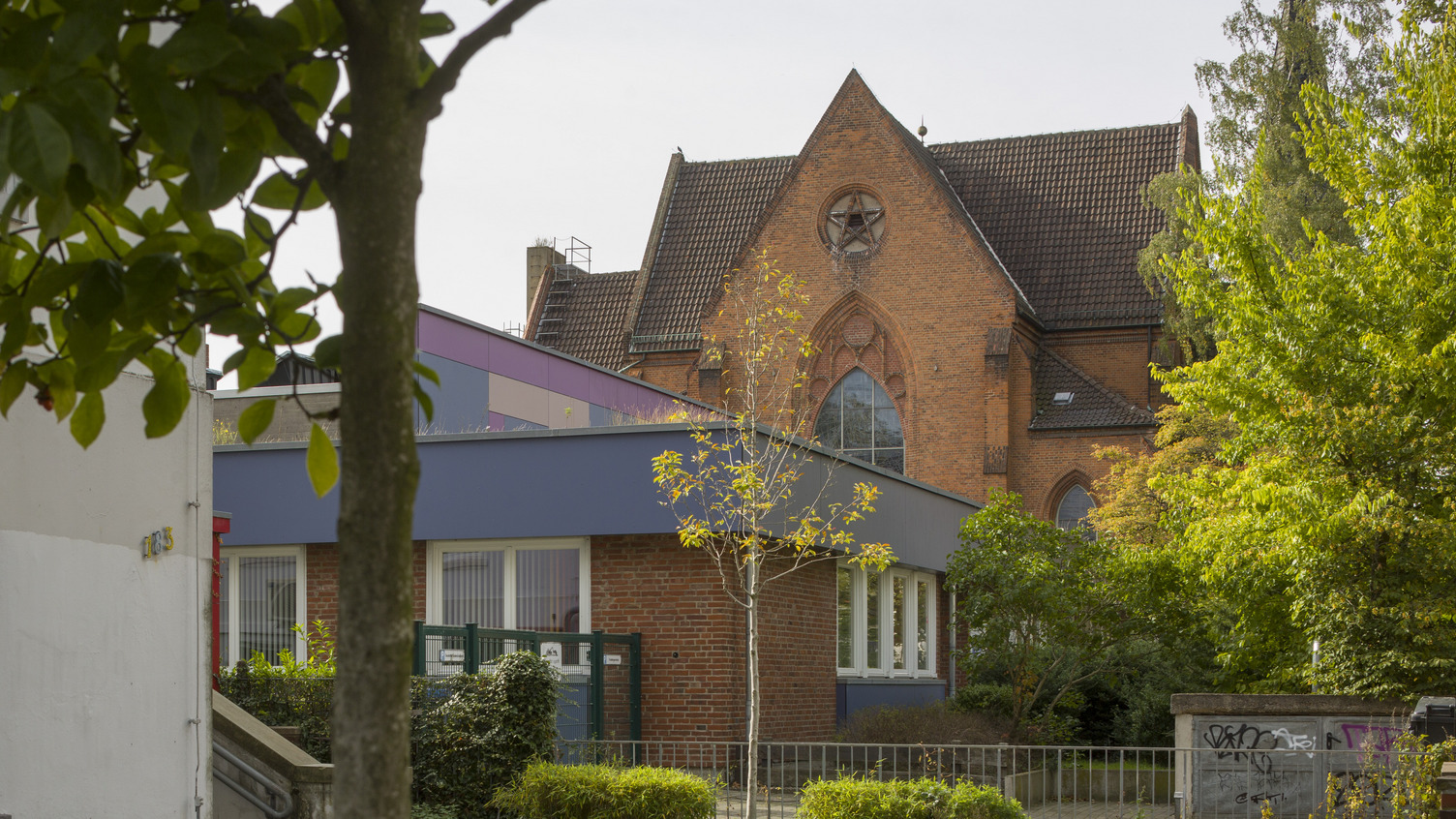 Ein rotes Backsteingebäude mit einem hohen, schrägen Dach und einem großen Fenster ist im Hintergrund sichtbar. Im Vordergrund stehen einige Bäume und ein kleiner Gartenbereich. Der Himmel ist klar, und die Umgebung wirkt ruhig und gepflegt.