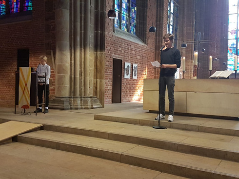 Man sieht wie zwei Jugendliche vorne in einer Kirche zur Gemeinde sprechen. Einer steht am Lesepult, einer vor dem Altar.