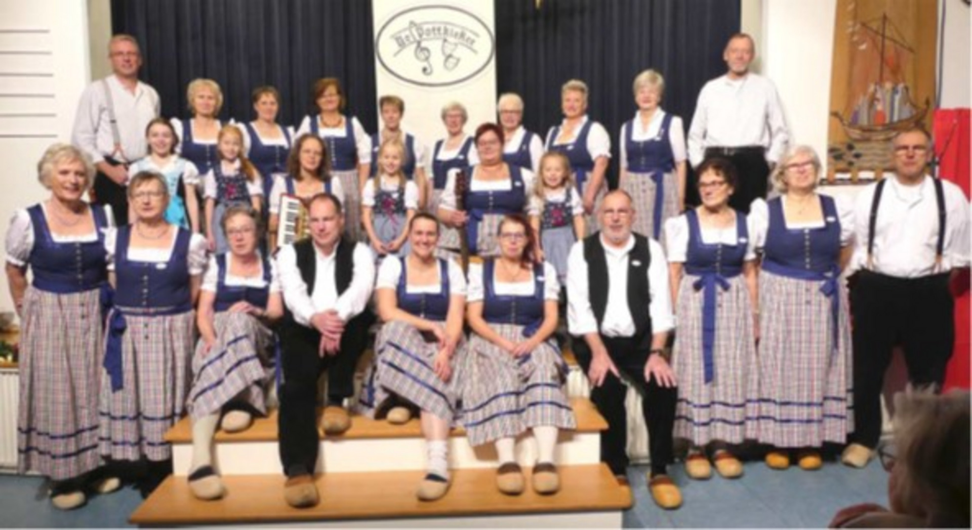 Eine Gruppe von 28 Personen in traditionellen Trachten steht und sitzt auf einer Treppe. Sie tragen meist blauen und karierten Stoff, dazu Holzschuhe. Im Hintergrund ist ein Banner mit dem Schriftzug „Heimatklänge“ zu sehen. Die Stimmung wirkt freundlich und gemeinschaftlich.