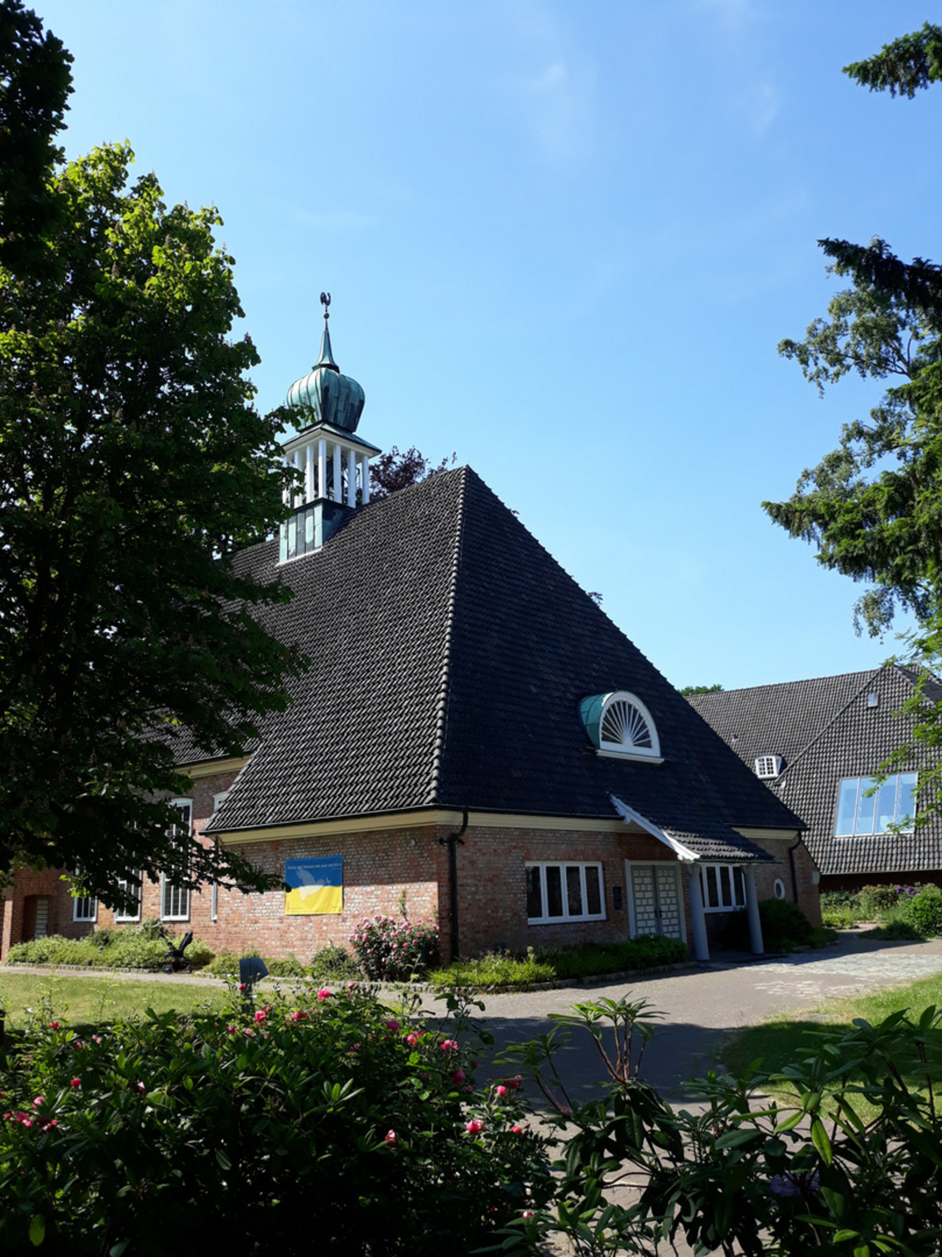 Ein Gebäude mit einer markanten, geneigten Dachform und einem Turm im Hintergrund. An der Fassade hängt eine Flagge, die die Farben der Ukraine zeigt. Die Umgebung ist grün mit Bäumen und blühenden Pflanzen. Der Himmel ist blau und klar.