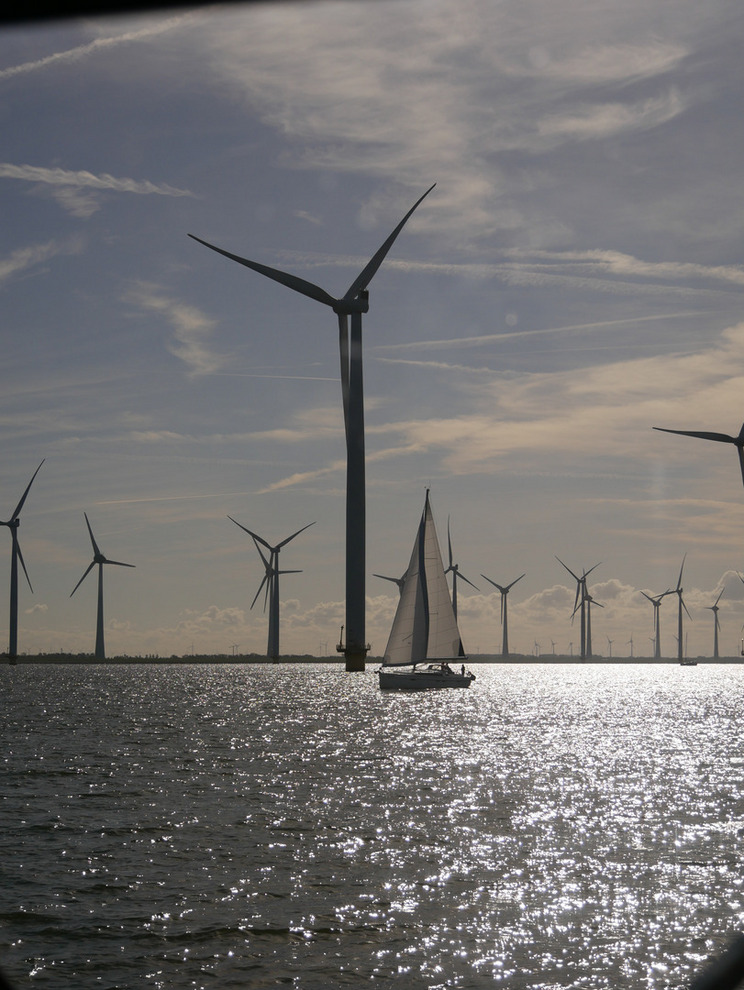 Ein Segelboot fährt über das glitzernde Ijsselmeer, umgeben von mehreren Windkraftanlagen, die sich gegen den Himmel abheben. Die Szene zeigt eine harmonische Verbindung von Natur und nachhaltiger Energieerzeugung.