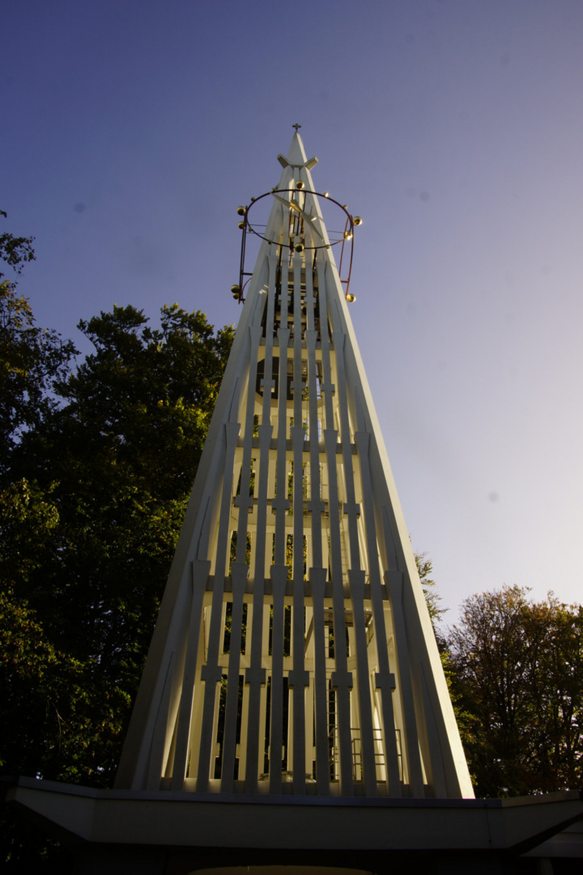 Ein schmaler, spitzer Turm aus weißen vertikalen Balken ragt gegen den klaren Himmel. An der Spitze befindet sich eine dekorative metallene Struktur. Um den Turm herum sind grüne Bäume zu sehen, die eine natürliche Umgebung schaffen.