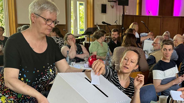 Das Bild zeigt eine Abstimmung im Kirchentag der Bremischen Evangelischen Kirche. Eine Frau steckt ihren Stimmzettel in eine Wahlurne.