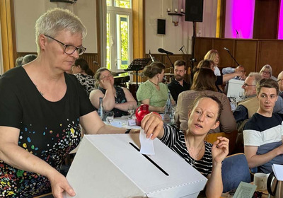 Das Bild zeigt eine Abstimmung im Kirchentag der Bremischen Evangelischen Kirche. Eine Frau steckt ihren Stimmzettel in eine Wahlurne.