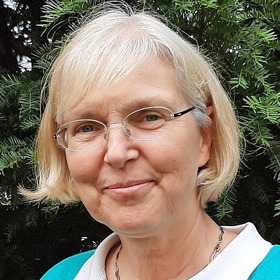 Eine lächelnde ältere Frau mit blonden, schulterlangen Haaren und Brille steht vor einem grünen Hintergrund. Sie trägt ein Oberteil in Türkis und Weiß und hat eine freundliche Ausstrahlung.
