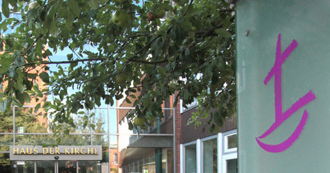 Das Bild zeigt den Eingang zum "Haus der Kirche". Die Glasfront ist modern und einladend, umgeben von Bäumen und einer kleinen Parkfläche für Fahrräder.