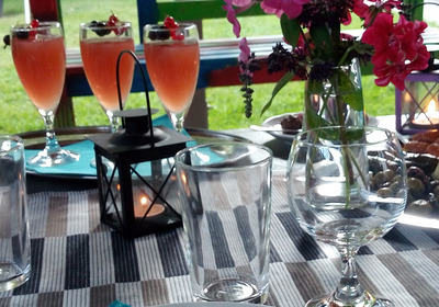 Der Tisch ist festlich gedeckt mit bunten Getränken in Gläsern, einer Vase mit bunten Blumen und einem Kerzenleuchter. Es gibt Besteck, Teller und Servietten in kräftigen Farben. Der Hintergrund zeigt eine grüne Wiese, die eine einladende Atmosphäre schafft.