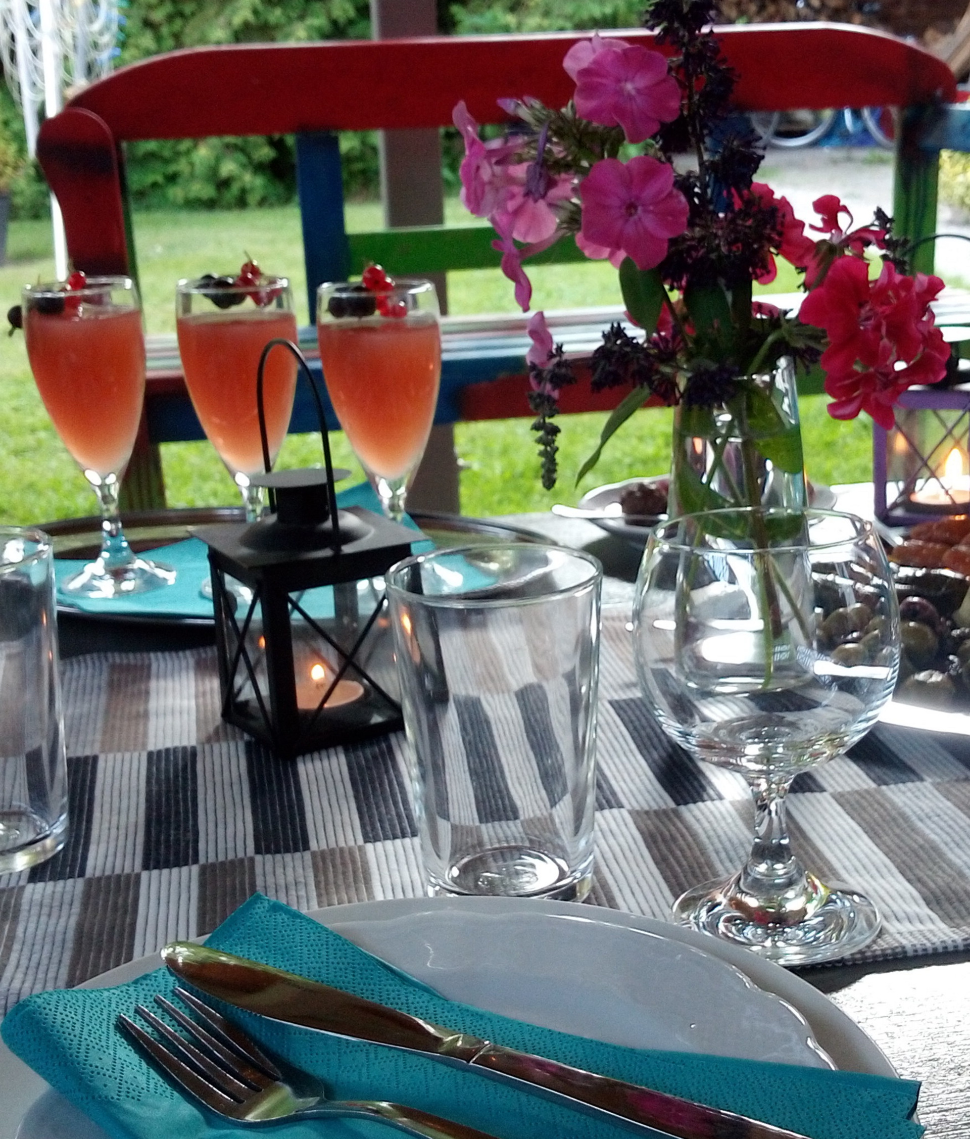 Der Tisch ist festlich gedeckt mit bunten Getränken in Gläsern, einer Vase mit bunten Blumen und einem Kerzenleuchter. Es gibt Besteck, Teller und Servietten in kräftigen Farben. Der Hintergrund zeigt eine grüne Wiese, die eine einladende Atmosphäre schafft.