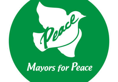 Rundes Logo in grün mit einer weißen Taube, die Frieden symbolisiert. Über der Taube steht das Wort "Peace" und darunter die Worte "Mayors for Peace". Das Design vermittelt eine Botschaft des Friedens und der Einheit.