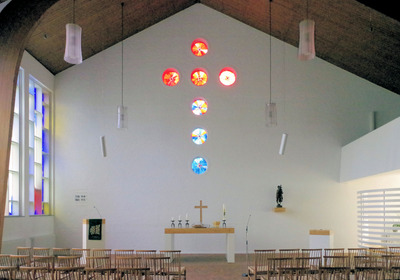 Das Bild zeigt das Innere einer modernen Kirche mit einer Wand, an der ein großes, buntes Fensterkreuz hängt. Darunter steht ein Altar mit einem Holzkreuz und Kerzen. Holzstühle sind in der Mitte des Raumes angeordnet. Die Wände sind hell und die Decke hat eine gewölbte Form.