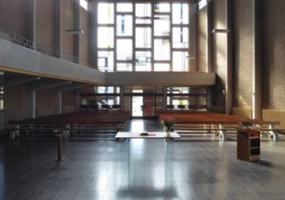 Eine schlichte Kirchenarchitektur mit langen Holzbänken, die auf einen zentralen Altar aus hellem Holz ausgerichtet sind. Große Fenster lassen Tageslicht herein, was den Raum erhellt. Der Bereich wirkt geräumig und ruhig, ideal für Andachten und Gebete.