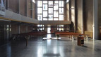Eine schlichte Kirchenarchitektur mit langen Holzbänken, die auf einen zentralen Altar aus hellem Holz ausgerichtet sind. Große Fenster lassen Tageslicht herein, was den Raum erhellt. Der Bereich wirkt geräumig und ruhig, ideal für Andachten und Gebete.