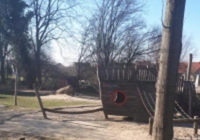 Ein Spielplatz mit einem großen, holzfarbenen Piratenschiff als Hauptattraktion. Das Schiff hat ein rundes Fenster und Seile zum Klettern. Im Hintergrund sind Bäume und ein klarer Himmel zu sehen. Der Boden ist mit Sand und Holzstücken gestaltet.