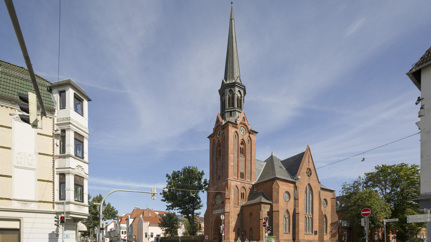 Ein historisches, rotes Backsteingebäude mit einem hohen, spitzen Turm und Zifferblättern an der Fassade. Es steht an einer Straßenecke, umgeben von Wohngebäuden und Bäumen unter einem klaren blauen Himmel. Verkehrsampeln und Straßenschilder sind ebenfalls sichtbar.
