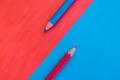Zwei Buntstifte liegen auf farbigem Papier. Der obere Stift ist blau und der untere Stift ist rot, wobei die Spitzen auf einer blauen und rot-orange-farbenen Fläche positioniert sind.