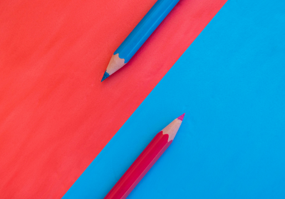 Zwei Buntstifte liegen auf farbigem Papier. Der obere Stift ist blau und der untere Stift ist rot, wobei die Spitzen auf einer blauen und rot-orange-farbenen Fläche positioniert sind.