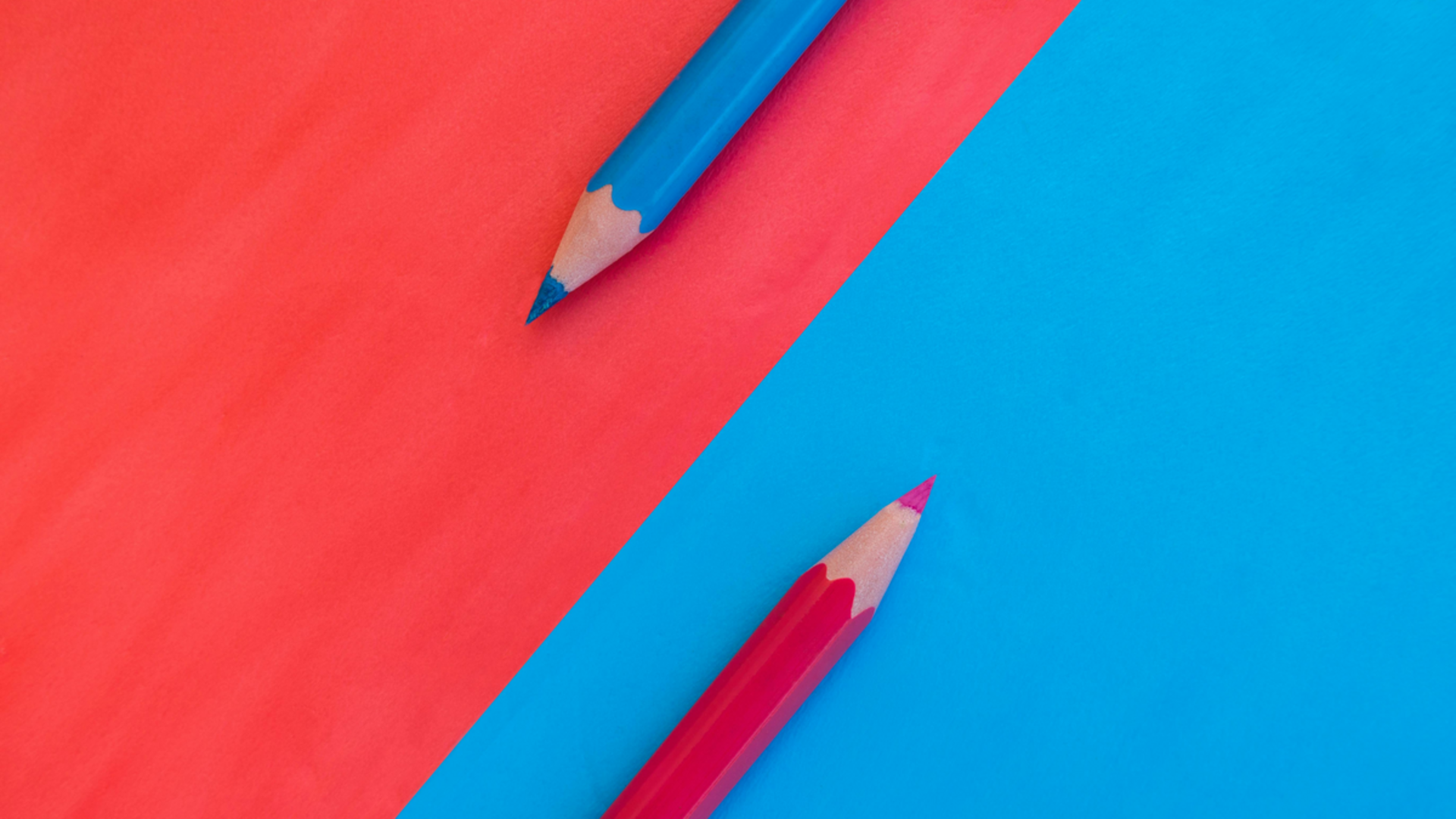 Zwei Buntstifte liegen auf farbigem Papier. Der obere Stift ist blau und der untere Stift ist rot, wobei die Spitzen auf einer blauen und rot-orange-farbenen Fläche positioniert sind.