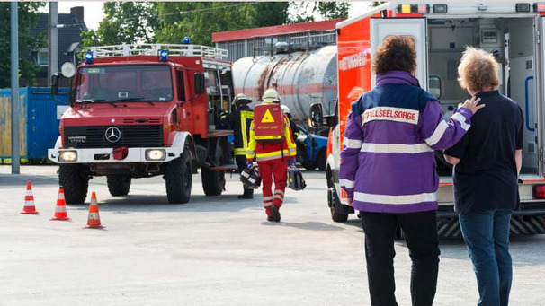 Das Foto zeigt einen Unglücksort. Im Hintergrund ein Feuerwehrfahrzeug, im Vordergrund ein Notfallseelsorge im Gespräch.