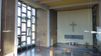 Ein Blick in den Innenraum einer modernen Kirche. An der Wand hängt ein großes Kreuz, und am Altar stehen Kerzen und eine Gedenktafel. Die großzügigen Fenster lassen viel Licht herein und zeigen ein schlichtes, aber einladendes Design.