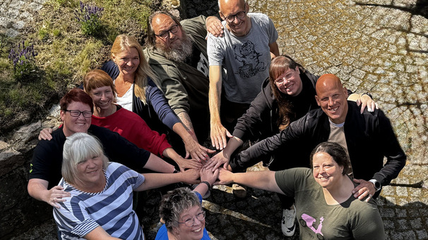 Eine Gruppe von zehn Personen steht im Freien auf einem Pflastersteinteppich. Sie lächeln und legen ihre Hände in die Mitte einer sich bildenden Gruppe von Händen. Die Atmosphäre ist freundlich und gemeinschaftlich.