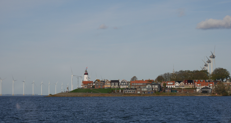 Urk Ein Blick auf eine Küstenlandschaft mit einem Leuchtturm und mehreren farbenfrohen Häusern. Im Hintergrund stehen Windkraftanlagen, die zur erneuerbaren Energieerzeugung beitragen. Das Wasser im Vordergrund reflektiert den klaren Himmel.