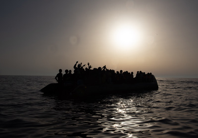 Das Foto zeigt ein völlig überladenes Flüchtlingsboot aus dem Meer. Es ist dunkel, im Hintergrund eine fahle Sonne