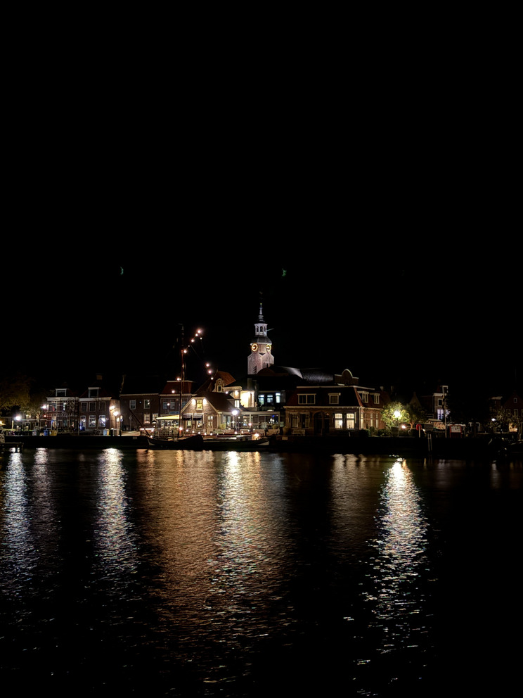 Blockzijl bei Nacht Der nächtlich beleuchtet Hafen Blockzijl, mit Gebäuden am Wasser. Reflektionen des Lichts auf der ruhigen Wasseroberfläche sind sichtbar. Im Hintergrund ragt ein Kirchturm in den Himmel, umgeben von der Atmosphäre einer ruhigen, klein-städtischen Umgebung.