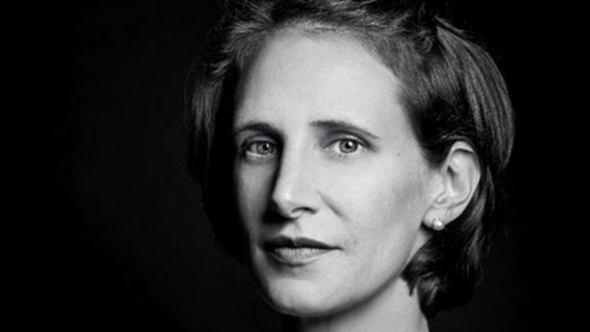 Porträt einer Frau mit kurzen Haaren, klaren Gesichtszügen und einem neutralen Ausdruck. Sie trägt Ohrringe und schaut direkt in die Kamera. Der Hintergrund ist dunkel, was den Fokus auf ihr Gesicht legt.
