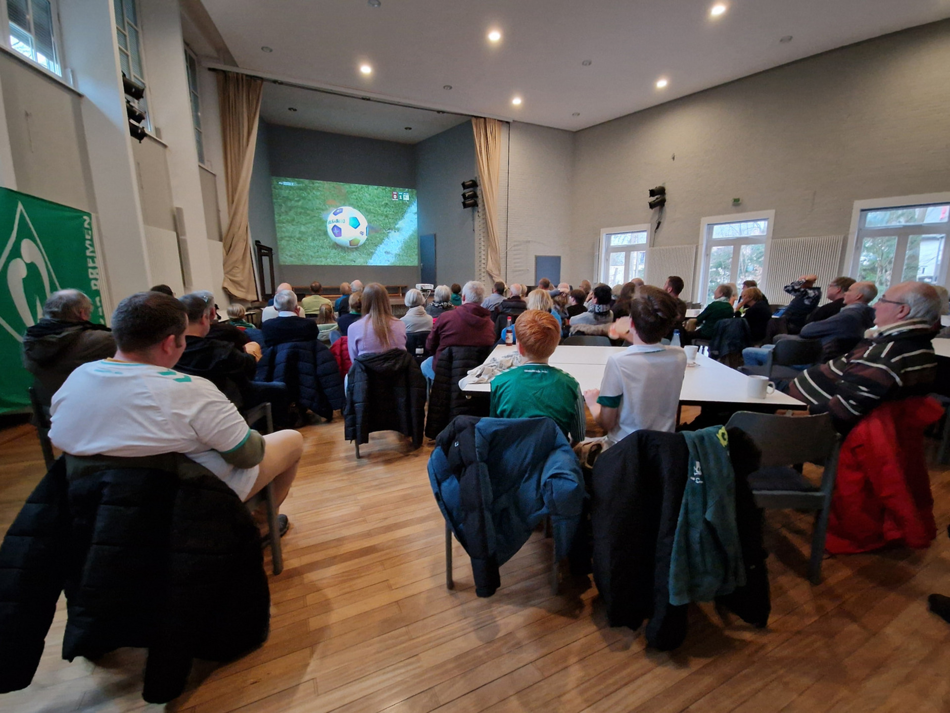 Eine Gruppe von Menschen sitzt in einem Raum und schaut auf eine große Leinwand, auf der ein Fußballspiel übertragen wird. Die Zuschauer tragen unterschiedliche Kleidung, darunter einige in Teamfarben. Im Hintergrund ist eine grüne Fahne sichtbar, die möglicherweise mit einem Fußballverein verbunden ist.