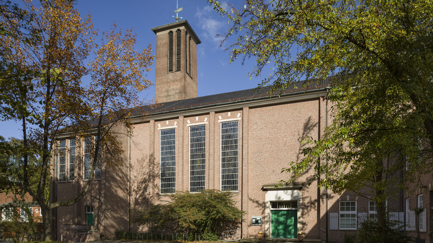 Das Bild zeigt eine Backsteinkirche mit großen Fenstern und einem schmalen Turm. Die Fassade ist von Bäumen umgeben. Ein grünes Eingangstor ist gut sichtbar, und der Himmel ist blau mit wenigen Wolken.