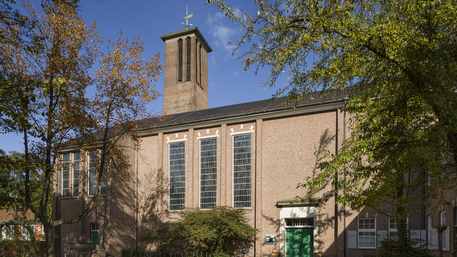 Das Bild zeigt eine Backsteinkirche mit großen Fenstern und einem schmalen Turm. Die Fassade ist von Bäumen umgeben. Ein grünes Eingangstor ist gut sichtbar, und der Himmel ist blau mit wenigen Wolken.