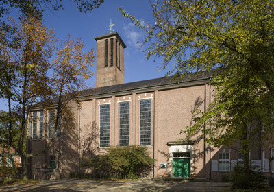 Das Bild zeigt eine Backsteinkirche mit großen Fenstern und einem schmalen Turm. Die Fassade ist von Bäumen umgeben. Ein grünes Eingangstor ist gut sichtbar, und der Himmel ist blau mit wenigen Wolken.
